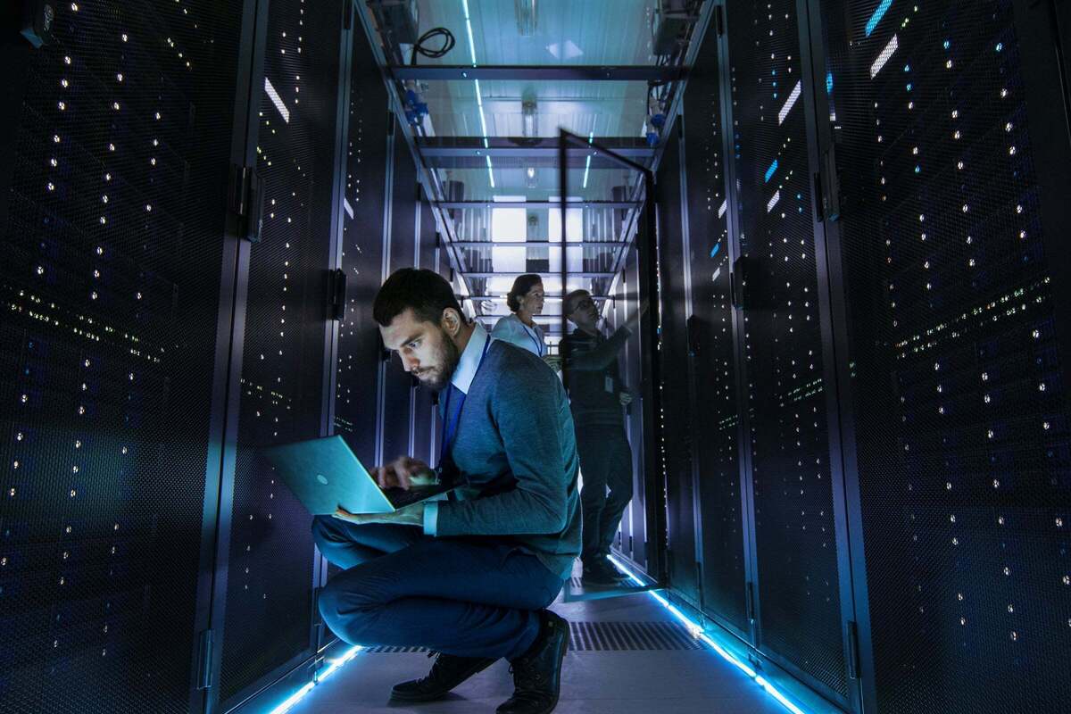 An IT technician works on laptop in data center, with other IT staff in the background.